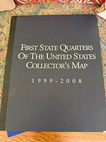 Closed collector's binder/map titled 'First State Quarters of the United States Collector's Map 1999-2008' in blue hardcover with black spine.