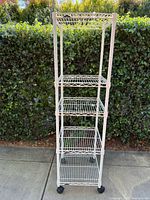 Front view of a white metal shelving unit with four wire shelves and caster wheels at the base. Background is greenery.