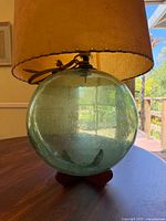 Front angle showing large green antique glass float, electrical wiring atop, mounted on wooden tripod base with attached beige fabric lamp shade.