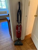 Full view of red Miele upright vacuum cleaner with black hose and silver handle, standing against wall