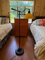 Full view of black metal floor lamp with round base and yellow dome-shaped shade placed between two beds in a room with wood-paneled walls, showing overall condition and design.