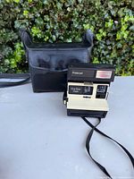 Front and front-left angled view of the Polaroid OneStep Flash instant camera showing the cream and black color scheme, flash unit, and included black carrying case in background.