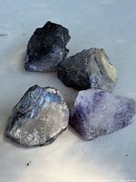 Four raw fluorite crystals in natural rough form on a light surface, showing color variations including purple and greenish-gray.