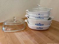 Stacked set of four white Corningware baking dishes with blue cornflower design and two clear glass lids on wooden countertop.