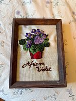 Frontal view of vintage crewel embroidery showing purple African Violet flowers, green leaves, red pot, and embroidered text on cream fabric in a wooden frame.