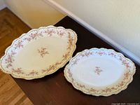 Two oval serving platters with pink rose floral pattern and gold trim, displayed on a wooden surface against a beige wall.