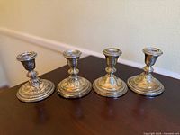Four vintage Empire sterling silver weighted candlestick holders on a dark wood surface, showing front view with ornate embossed designs.