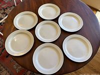 Top view of seven cream-colored enamel dinner plates with black rims arranged in a circle on a wooden table, showing minor surface wear.