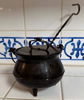 Front view of the black cast iron cauldron with lid and side handles on tiled surface with blue decorative backsplash.