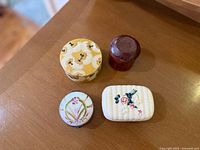 Four vintage handmade powder and trinket boxes on a wooden surface, showing various shapes, materials and floral designs.