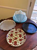 Four vintage mid-century platter trays grouped on wooden table: blue round tray with mesh cover, cream scalloped edged tray, black tray, and cream tray with red paisley pattern.