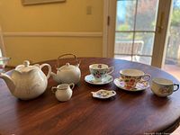 Wide shot showing two porcelain teapots, a creamer, three teacups with saucers, and a small decorative plate on a wooden table in front of a window.