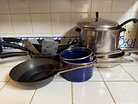 Seven vintage cookware items on a kitchen counter including stainless steel pot with lid, blue enamel saucepans, cast iron skillet, and frying pans.