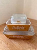 Four vintage Pyrex refrigerator dishes stacked, showing two butter yellow rectangular dishes with clear lids and two smaller white opaque dishes, all with Butterfly Gold floral gold pattern.