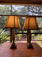 Photo of two vintage table lamps placed on a wooden surface near a window, showing fabric shades and decorative wooden bases.