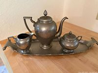 Full view of the vintage metal tea set on a wooden surface, showing teapot, creamer, sugar bowl, and tray with patina finish.