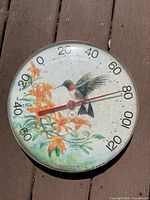 Photo of vintage outdoor jumbo dial thermometer with hummingbird and flower design on wooden background, shows dial from -40 to 120 degrees Fahrenheit.