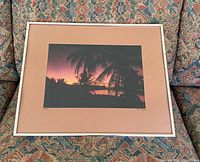 Photo showing one framed vintage tropical sunset art print with palm tree silhouettes, peach matte, and white frame, lying on patterned fabric.