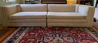 Full view of the vintage two-section beige sofa featuring two seat and back cushions on each section, curved arms, and wooden feet, set against a decorative rug and wooden floor.