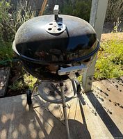 Photo of the vintage Weber charcoal BBQ pit outdoors showing the grill's rounded black lid with top vent, metal legs and wheels on a patio.