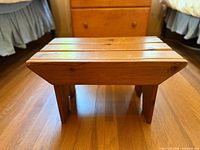 Front left angle view of the small wooden step stool showing its slatted top and legs on wood floor.
