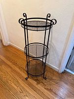Full view of the black wrought iron 3-tiered stand on a hardwood floor against a wall.