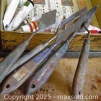 Four palette knives on aged wooden surface with oil paint tubes partially visible in background