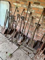 Group of long-handled shovels, rakes and cultivators leaning against brick wall