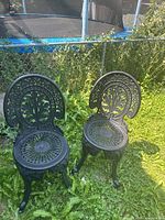 Two black cast iron chairs on grass with ornate backs and round seats