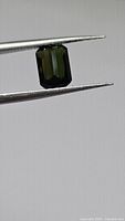 Close-up image of a deep forest green, octagon cut tourmaline gemstone held by gemstone tweezers against a plain background, showing facets and clarity.