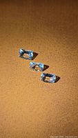 Three small cushion cut aquamarine gemstones displayed on a brown textured surface. The stones show light blue color and are faceted.
