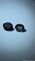 Pair of natural blue-green oval cut sapphire gemstones placed on a white background, showing color and clarity.
