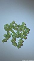 Photo showing about 40 small rough demantoid garnet crystals with greenish-yellow hues, scattered on a white background, under good lighting to show color and crystal form.