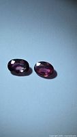 Photo showing two oval cut sapphires in deep pink purple shades on a plain light background, highlighting their cut and color.