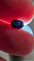Close-up image showing oval cut, deep blue spinel gemstone held between fingers with visible facets and light reflection.
