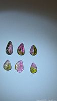Six leaf-shaped watermelon tourmaline gemstones arranged on a plain surface showing their natural bi-color pink and green hues with carved leaf vein details.