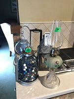 Group showing blue mosaic oil lamp, green floral hurricane lamp, clear glass hanging lantern, black metal mesh lantern