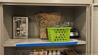 Shelf view with OXO cookie press box, green basket of attachments, KitchenAid processor base