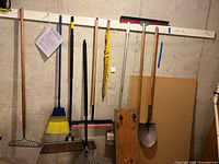 Various long-handled tools hanging on wall: rake, push broom, shop broom, mop handle, squeegee, shovel