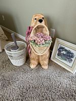 Front view of ceramic dog figurine, wicker wastebasket, toilet plunger inside, framed print leaning against wall