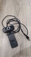Front view of USB webcam showing lens, microphone grille, attached USB cable and clip mount