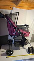 Side view of burgundy Pet Gear pet stroller showing frame, wheels, mesh cabin, storage basket