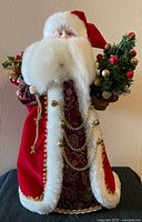 Front view of plush standing Santa figurine showing red velveteen coat, white faux fur trim, gold chains, and miniature Christmas tree in right hand