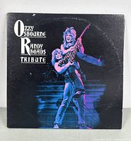 Front cover of 'Ozzy Osbourne & Randy Rhoads Tribute' double vinyl record album showing Ozzy Osbourne lifting Randy Rhoads on stage with electric guitar under purple lighting.
