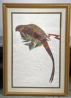Framed artwork showing a long-tailed bird holding colorful leaves on an embossed white background, gold ornamental frame visible.