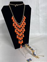 Necklace displayed on black jewelry stand showing cascading orange teardrop beads on gold-tone chain, with bracelet beside on clear stand showing blue stones.