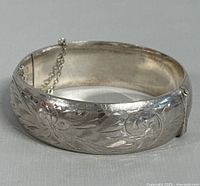 Close-up photo showing the hand-engraved floral design and the safety chain on the hinged silver bangle.