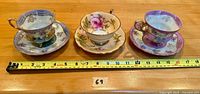 All three footed teacup and saucer sets displayed side by side on a wooden surface with a measuring tape indicating approximate size.