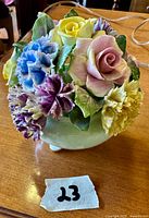 Close-up of delicate floral arrangement with pink rose, yellow rose, purple and blue carnations and green leaves in a pedestal bowl.