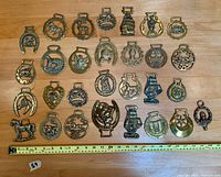 Photo showing 29 vintage brass horse brasses arranged in 4 rows on wooden surface with measuring tape for scale.